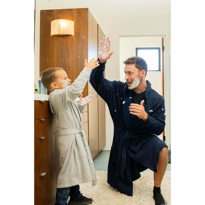 A fun moment of a man in The DudeRobe - Men's Luxury Hooded Bathrobe playing with a child in a bathroom.