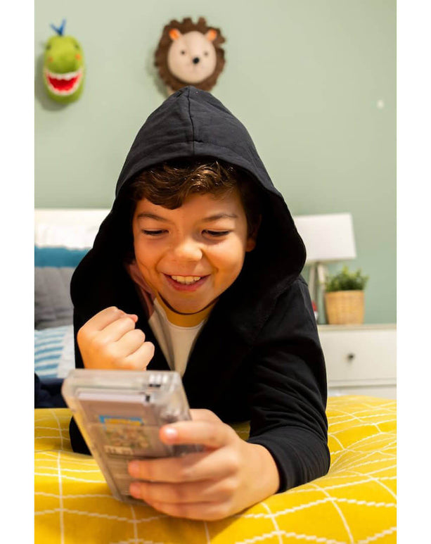Smiling boy in a hooded black bathrobe playing with a video game on a cozy bed.