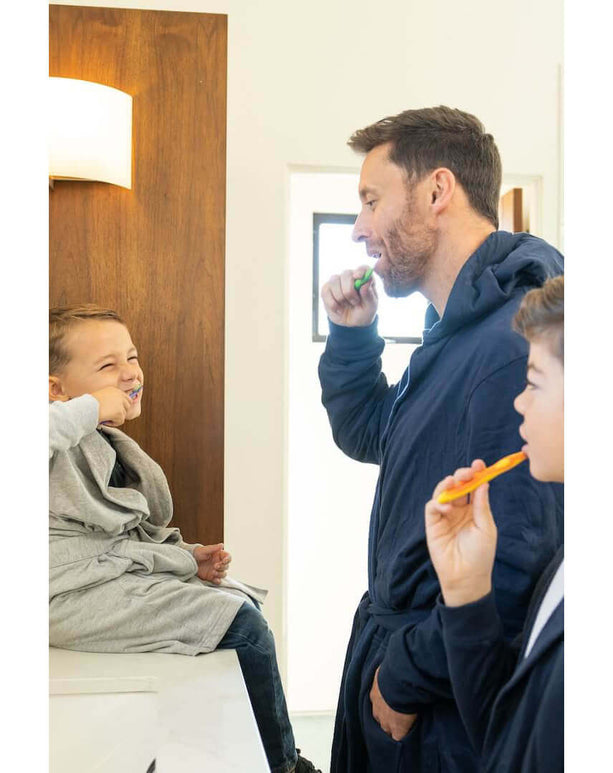 A father and two boys brush their teeth while wearing luxury kids hooded bathrobes in a bright bathroom.