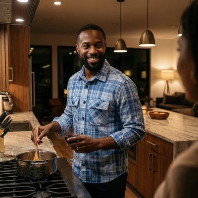 A man wearing The 1-Second Flannel magnetic button shirt cooks in a modern kitchen, smiling and enjoying a glass of wine.